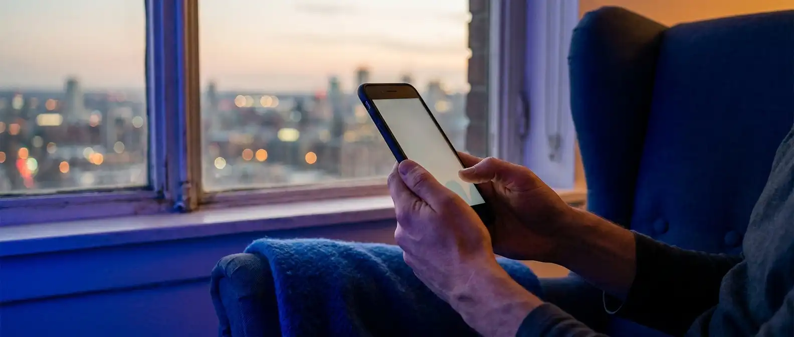 Someone seated in an armchair at dusk, holding a phone, with city lights out of focus beyond the window